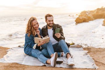 Image of young caucasian couple using cellphone while walking by seaside