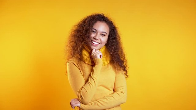 Pretty young girl with curly hair standing on yellow studio background, smiling and flirting. Cute portrait of mixed race woman