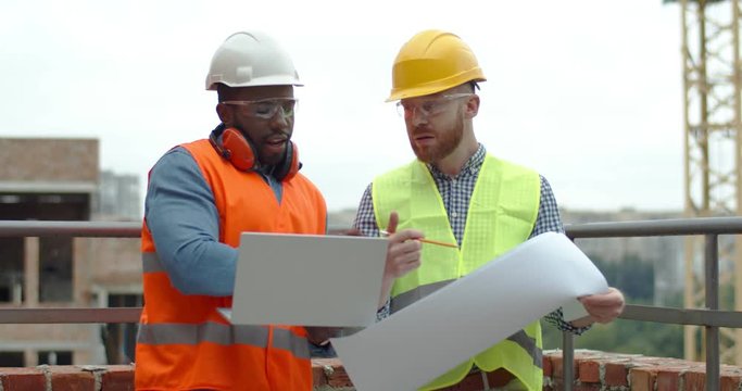African American young handsome contractor with laptop computer in hands talking and discussing construction with Caucasian builder with a building plan in hands. Outdoor.
