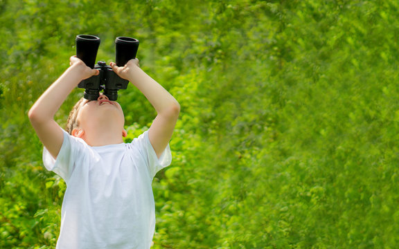 banner size. curious child boy in white t-shirt looking through binoculars in nature green landscape. Explore and discover wildlife concept. kid adventure. copy space for text.