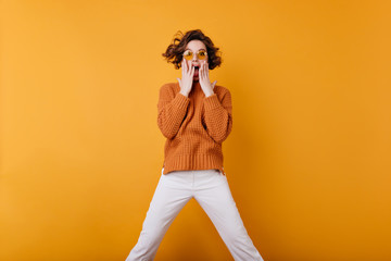 Gorgeous emotional woman in trendy yellow sunglasses expressing amazement. Indoor portrait of brown-haired girl wears white pants and cozy sweater.