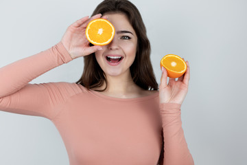 young woman holding one half of oranges near her eye and another near shoulder smiling standing on...