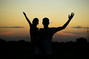 Fototapeta na wymiar Silhouettes of father and daughter on his shoulders with hands up having fun, against sunset sky. Parenthood, family activities, vacation, support and love themes