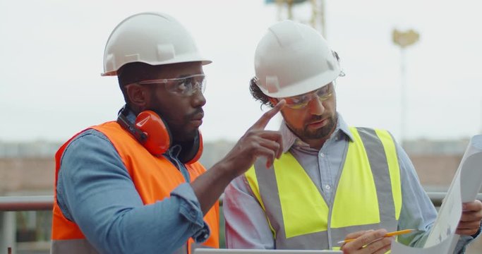 Close up of the two mixed-races male constructors in goggles and hardhats having a talk and discussion about building cunstruction plan. Outside.