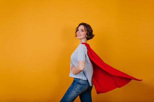 Smiling supergirl in jeans standing in confident pose on yellow background. Indoor photo of curly lady preparing for new year carnival.