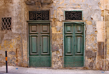 Two derelict green doors with ornate grills above them in a crumling limestone wall in Sliema, Malta