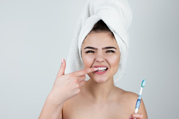 young beautiful woman wearing white towel on her head holding tooth brush with toothpaste touching her tooth with finger on isolated white background, morning routine