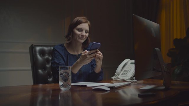 Young businesswoman working in night office. Woman laughing looking at smartphone sitting at dest with monitor. 