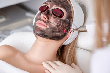 Close-up of cosmetologist's hand making hardware carbon peeling by young pretty customer woman in safety glasses headdress lying on couch in beauty salon. Facial cleaning concept. Cosmetology Services
