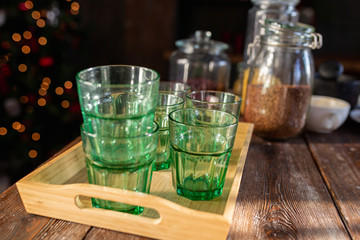 Empty green glass glasses on a wooden tray on a wooden table in the sunlight against the background of kitchen supplies.