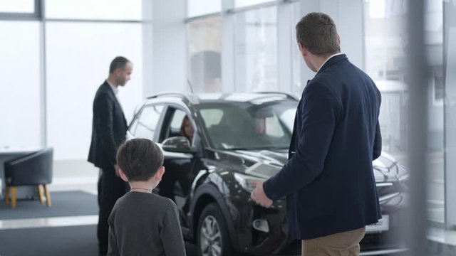 Back view of little brunette Caucasian boy talking with car dealer as his parents selecting new vehicle at the background. Successful family buying automobile in dealership.
