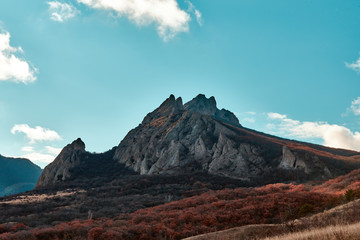 view of a rocky mountain at the foot of which is an autumn forest, the sky is blue with clouds