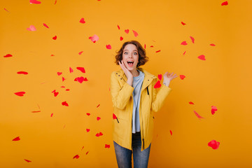 Excited white girl with light-brown wavy hair posing in valentine's day. Indoor photo of happy young woman standing under fallen hearts and laughing.
