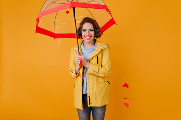 Smiling happy caucasian girl holding red parasol on yellow background. Indoor photo of graceful...