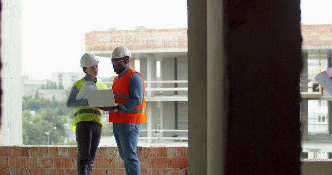 View from inside on the Caucasian couple of constructors, man and woman, standing at the terace outdoor and discussing drafts of the construction.