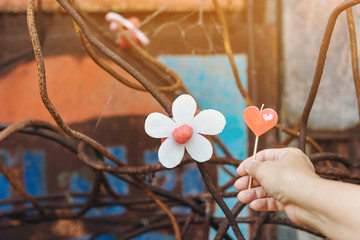 A red heart-shaped candle on woman's hand and an artificial flower made of white and red candles with bluerd image of rusty steel rod in the background.