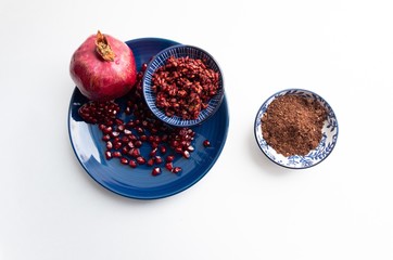 Flat lay of ground pomegranate seeds spice  in bowl on white background with garnet fruit and berries in blue plate