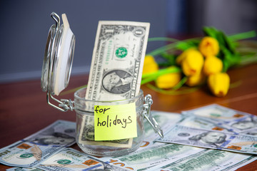 One Dollar bill in glass jar on wooden table with money background. Saving money for holidays concept