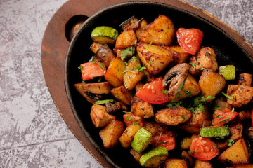 Delicious oven-baked potatoes with herbs and vegetables in a pan, rustic style. Potatoes fried, red cherry tomato, zucchini, onion on gray stone background.