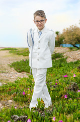 Full body portrait of boy with arms and legs crossed on the plants wearing a fashion white sailor suit for his communion