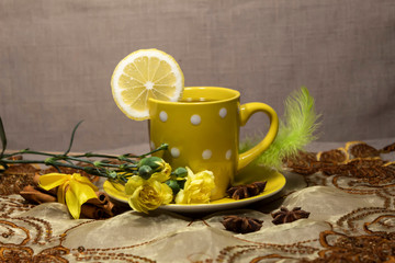 A cup of tea with lemon stands on the table. On the saucer are yellow carnation flowers. Nearby are sticks of cinnamon and star anise. Behind there is a beautiful green feather.