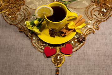 A cup of tea with lemon is on a bright oriental napkin. On a saucer lies star anise, cinnamon sticks and yellow clove flowers. In front are two red hearts.