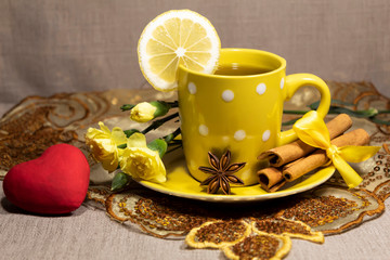 A cup of tea with lemon stands on the table. On a saucer are cinnamon sticks, star anise and yellow clove flowers. Nearby lies a red heart.