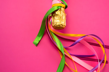 Champagne bottle neck in gold foil and colorful colored satin ribbons on bright pink background, flat lay top view. Concept of celebration and celebrating, birthday, congratulations, happiness, party 