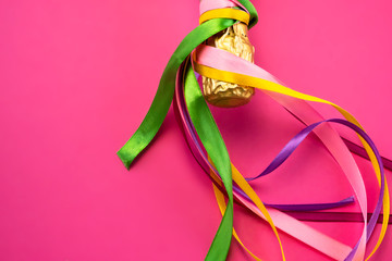 Champagne bottle neck in gold foil and colorful colored satin ribbons on bright pink background,...