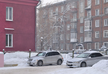 Fototapeta na wymiar Two cars in the courtyard of the city house. Two silver cars and two buildings. The road and lawns are covered with snow. Winter.