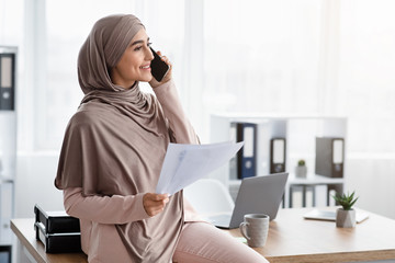 Millennial muslim businesswoman holding papers and talking on cellphone in office