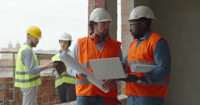 Multiethnic two men co-workers, builder with architect disputing about construction with laptop computer and plans in hands, man and woman constructors talking on the background.