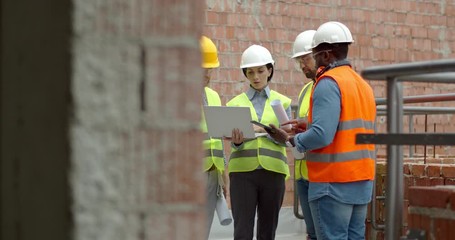 Caucasian woman architect in hardhat showing some plan of building to the male mixed-races workers while they have working meeting at the balcony on the construction site. - Powered by Adobe