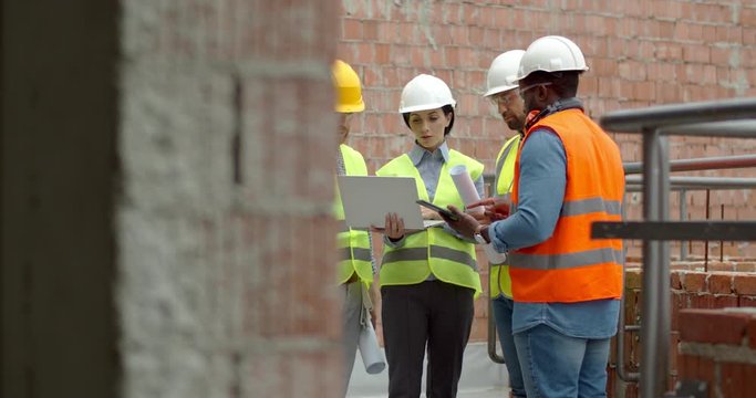Caucasian woman architect in hardhat showing some plan of building to the male mixed-races workers while they have working meeting at the balcony on the construction site.