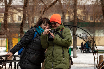 Two girls take pictures and walk in a city park on a sunny day in early spring. A girl photographer takes a picture of a model and then watches the photos together on the camera.
