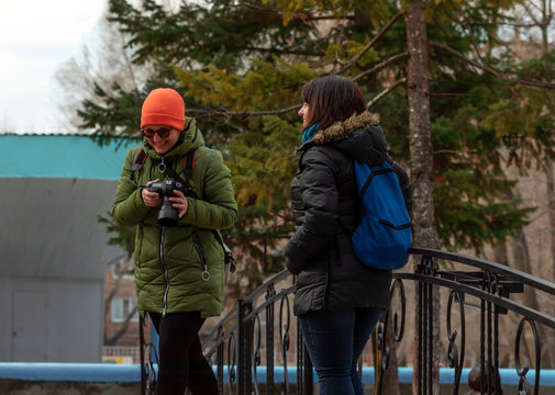 Two girls take pictures and walk in a city park on a sunny day in early spring. A girl photographer takes a picture of a model and then watches the photos together on the camera.