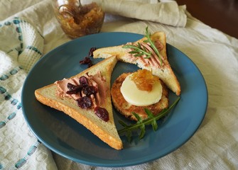 A generous toast breakfast with pate, cheese and onion marmalade on a blue plate, decorated with dried cranberries and fresh arugula.