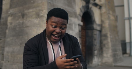 Obraz premium Young african guy making surprising face and looking at smartphone screen while standing outdoors. Millennial male looking extremely happy and satisfied of good news. Old city background.