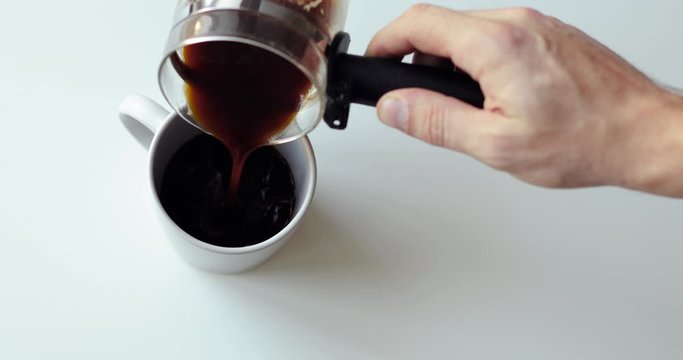 Preparing black coffee on white table. Barista is making fresh espresso coffee pouring it to cup from glass turk, top view, hand closeup. Beverage with caffeine for cheerfulness.