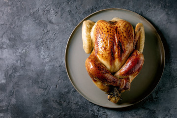 Whole baked chicken poultry on ceramic plate over black concrete background. Flat lay, space. Dinner menu