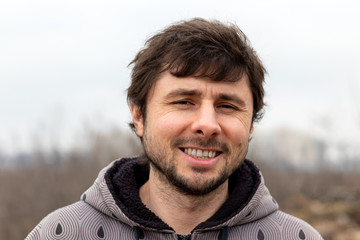 A handsome happy smiling bearded man outside against a blurred background. Man in a jacket with a hood