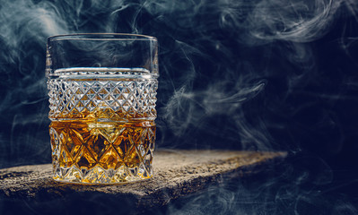 glass of whiskey with ice on a wooden table surrounded by smoke