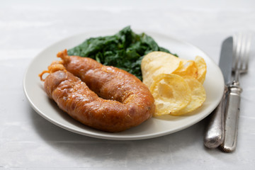 fried smoked sausage with potato chips and greens on white plate on ceramic background