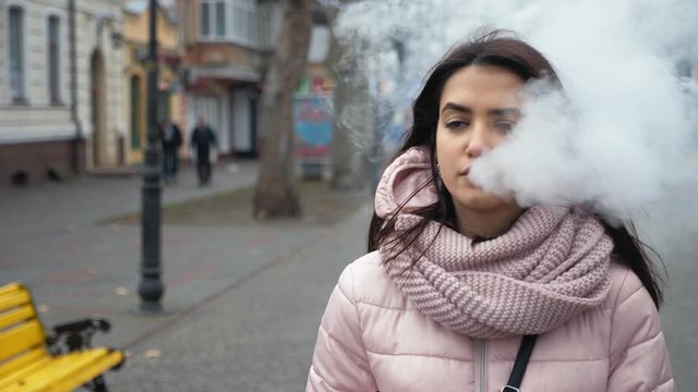 Beautiful calm girl walking through the street with e-cigarette and vaping, slow motion