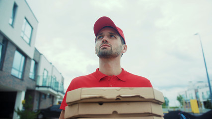 Worried pizza man carrying carton boxes walking in the street. Portrait of hurrying courier looking on watch with surprising expression searching for the right house in the courtyard.