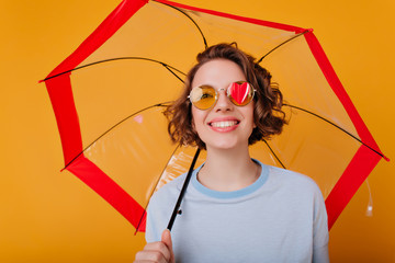 Studio shot of cute white lady isolated on yellow background with parasol in hand. Curly female model with interested smile posing under umbrella.