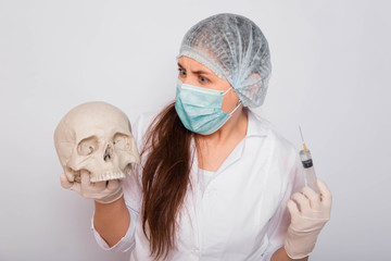 Female doctor in a white medical coat, mask, hat and gloves holds a human skull and a syringe with a vaccine in her hands.