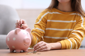 Woman putting coin into piggy bank at wooden table, closeup