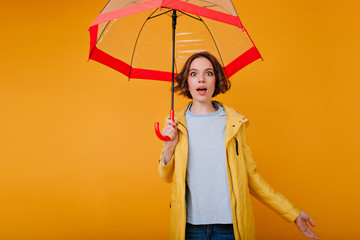 Ecstatic young caucasian woman in blue shirt and yellow coat expressing amazement. Indoor photo of curly dreamy girl having fun during photoshoot with parasol.