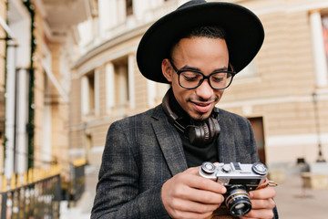 Smiling male photographer in headphones standing on city background. Outdoor photo of cheerful african young man in checkered jacket looking at camera.